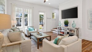 A living room with a ceiling fan, a TV mounted on the wall, and a bookshelf. - 30A Treetop Retreat • Shared Pool + Private Beach Access + Bikes | Pura Vida (Seacrest Beach)
