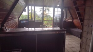 A wooden interior with a reception desk, a television, and a view of palm trees and mountains through large windows. - Beautiful chalet with panoramic views (Ubatuba)