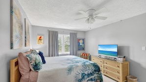 A bedroom with a bed, a television, a ceiling fan, and artwork on the walls.
