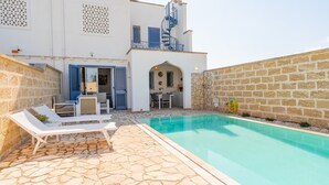 A poolside area with lounge chairs, a stone-tiled patio, and a building with a blue spiral staircase and shutters. - VILLINO LORENZO for Rent 3 min from Porto Cesareo with Pool (Boncore)