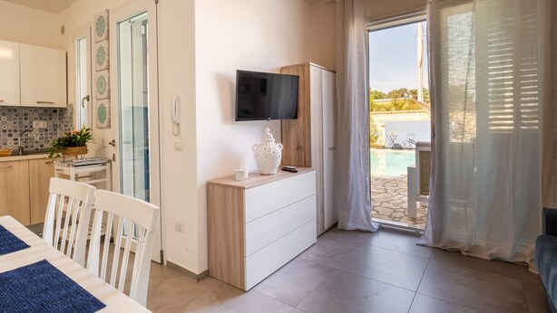 A modern room with a TV mounted on the wall, a wooden dresser, a dining table with chairs, and a view of an outdoor pool area. - VILLINO LORENZO for Rent 3 min from Porto Cesareo with Pool (Boncore)