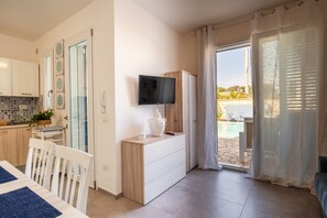 A modern room with a TV mounted on the wall, a wooden dresser, a dining table with chairs, and a view of an outdoor pool area. - VILLINO LORENZO for rent, 3 minutes from Porto Cesareo, with a pool (Boncore)