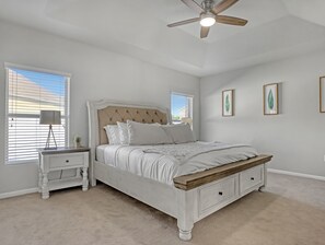 A bedroom with a bed, nightstand, ceiling fan, and large windows.