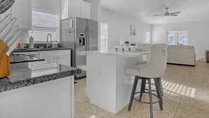 A modern kitchen with a central island, a refrigerator, and a dining area with a bar stool. - Melbourne Beach House (West Melbourne)