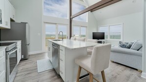 A modern kitchen with a central island, a dining area with a sofa, and a large window offering a view of the outdoors. - 5260-Shenanigan's, 3BR Soundfront Home | Gated Community Pool (Kill Devil Hills)