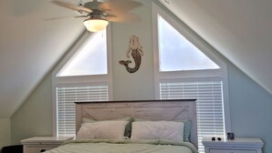 A bedroom with a sloped ceiling, a bed with a wooden headboard, two bedside tables, a ceiling fan, and two windows with blinds. - 5260-Shenanigan's, 3BR Soundfront Home | Gated Community Pool (Kill Devil Hills)
