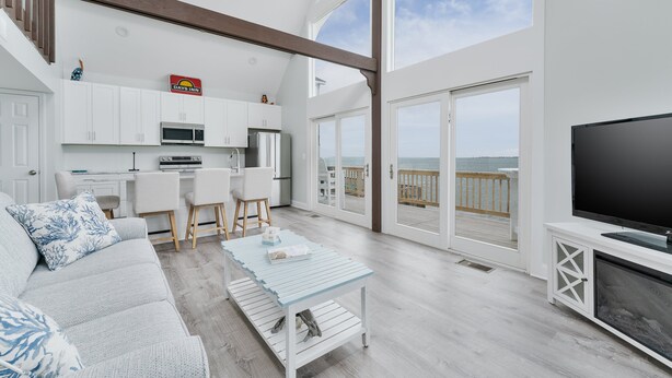 A modern living room with a sofa, coffee table, TV, and a balcony with a view. - 5260-Shenanigan's, 3BR Soundfront Home | Gated Community Pool (Kill Devil Hills)