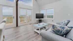 A spacious living area with a balcony, a flat-screen TV, and a light blue coffee table. - 5260-Shenanigan's, 3BR Soundfront Home | Gated Community Pool (Kill Devil Hills)