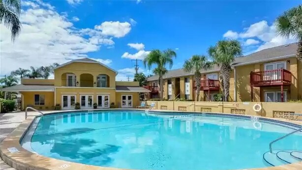 A swimming pool surrounded by a brick walkway, with a building featuring balconies and a yellow facade in the background. - Lovely 3 Bedroom Condo On Golf Course (Sarasota)