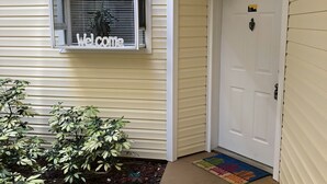 A house exterior with a 'Welcome' sign in the window, a white door, and a small garden area with plants and a colorful rug. - Precious Condo, 3 Blocks to Beach.. Happy Place! (Indialantic)