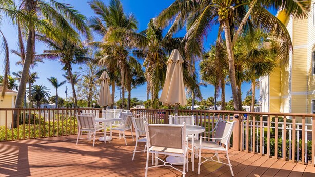 A wooden deck with white metal chairs and tables, surrounded by palm trees and a clear blue sky. - Precious Condo, 3 Blocks to Beach.. Happy Place! (Indialantic)