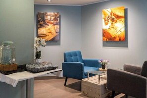 A modern room with a blue armchair, a glass coffee table, and a white table with a tray of glasses. There are two paintings on the walls, one depicting a kitchen scene and the other a workshop.