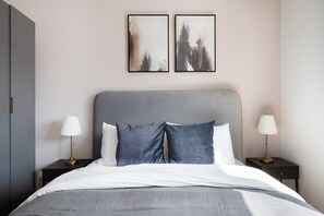 Un dormitorio moderno con cabecera gris, ropa de cama blanca, dos lámparas de noche y arte abstracto en la pared.