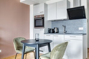 Uma cozinha compacta com micro-ondas, forno e cafeteira, uma pequena mesa redonda com duas cadeiras, e um copo.