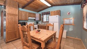 A wooden dining table with chairs, a kitchen with wooden cabinets, a refrigerator, and a clock on the wall. - Roadrunner - Historic Upper Canyon, Priv Hot Tub, Cozy (Ruidoso)