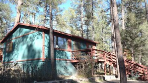 A rustic cabin with a wooden deck and a large window, surrounded by tall trees. - Roadrunner - Historic Upper Canyon, Priv Hot Tub, Cozy (Ruidoso)