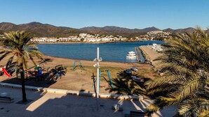 Ein Strand mit Spielplatz, Palmen und Blick auf eine Küstenstadt mit Booten und Bergen im Hintergrund.