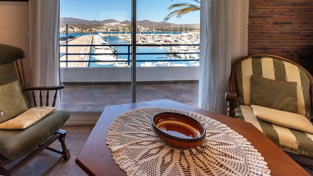 A room with a view of a marina, a wooden table with a bowl, two chairs, and a large window. - Port de Llançà 34 - Apartment with sea views (Llança)
