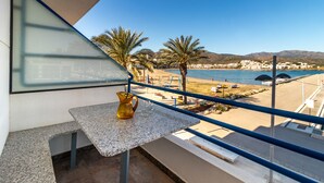 A balcony with a table, a pitcher, and a clear view of a beach, palm trees, and a mountainous landscape. - Port de Llançà 34 - Apartment with sea views (Llança)