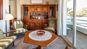 A room with a wooden table, two upholstered chairs, a cabinet with decorative plates, and a view of the outdoors. - Port de Llançà 34 - Apartment with sea views (Llança)