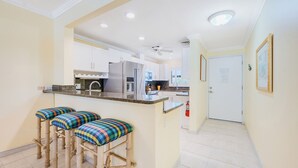 Une cuisine avec des armoires blanches, un comptoir en granit et un ventilateur de plafond. On y trouve trois tabourets dotés de coussins à carreaux.