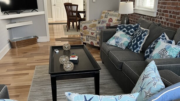 A living room with a grey sofa, a coffee table, and a TV on a stand. There is a door leading to another room and a window with blinds. - Adorable 1st floor apartment 2 blocks to the beach. (Ventnor City)