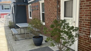 A building with a brick facade, a welcome sign, a covered patio with a table and benches, and potted plants. - Adorable 1st floor apartment 2 blocks to the beach. (Ventnor City)