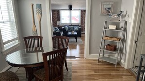 A dining area with a glass-top table and wooden chairs, a brick wall in the background, and a bookshelf with baskets. - Adorable 1st floor apartment 2 blocks to the beach. (Ventnor City)