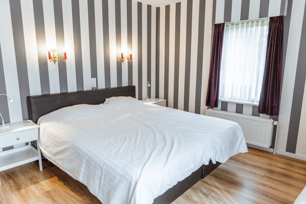 Un dormitorio ordenado con una cama grande, mesitas de noche, una lámpara de pared y una ventana con cortinas.