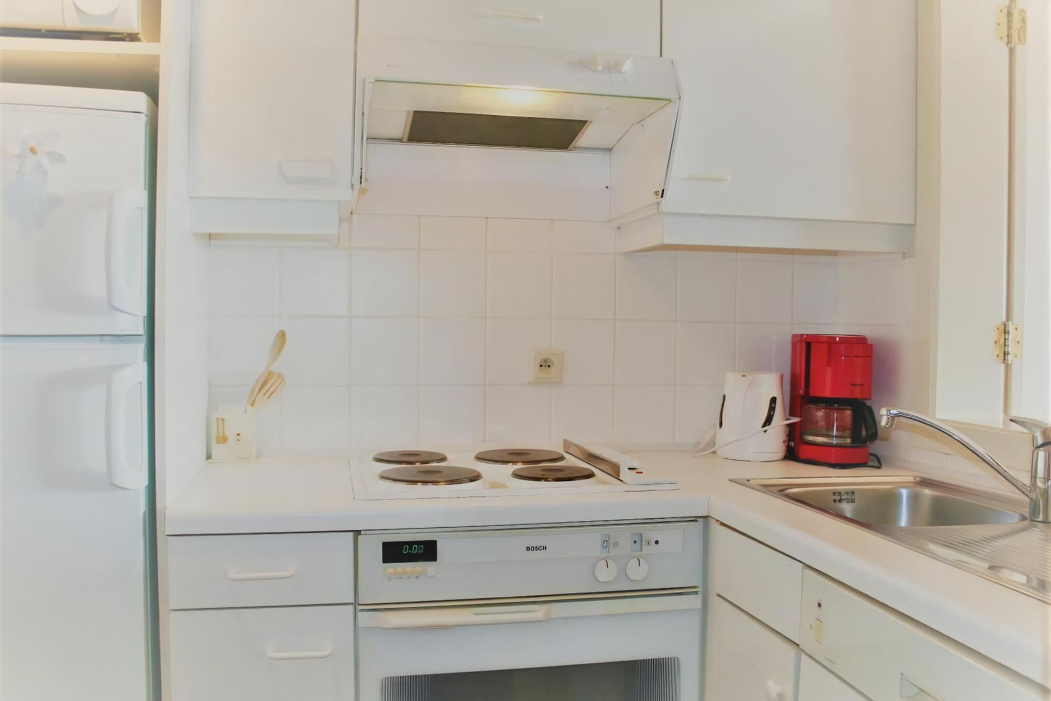 Una cocina con gabinetes blancos, una estufa Bosch, una cafetera roja y un lavamanos.