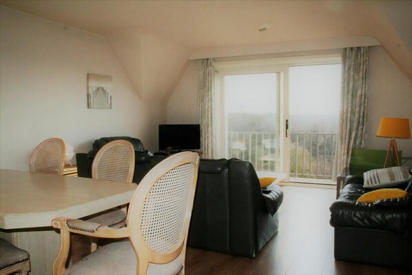 Ein Zimmer mit Esstisch, Stühlen, einer Couch, einem Fernseher und einem Balkon mit Vorhängen.