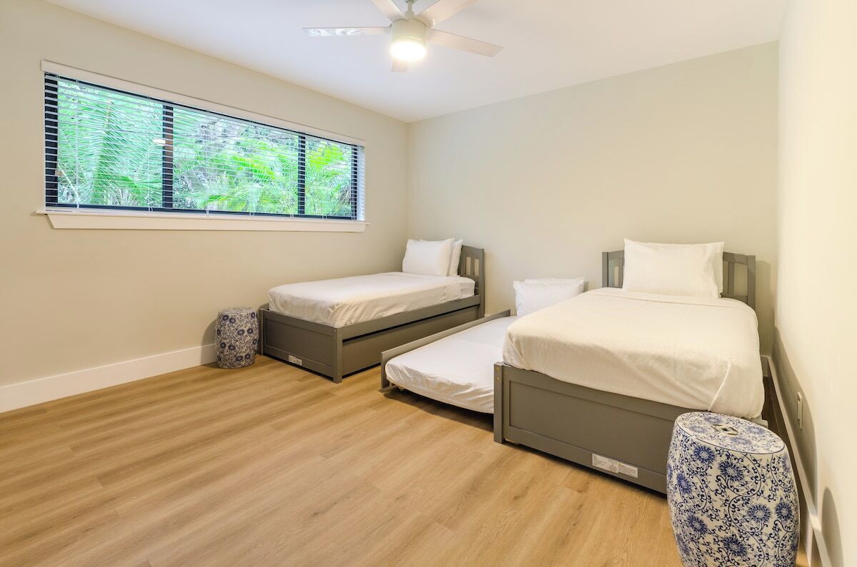 A bedroom with two single beds and a larger bed, a ceiling fan, a window with blinds, and a wooden floor.