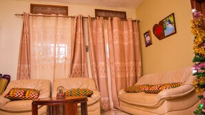 A living room with two beige leather sofas, patterned cushions, a wooden coffee table, and a window with patterned curtains. - 3 Bed House - Sleeps 8, Wifi, Parking, Pets (Kampala - Uganda)