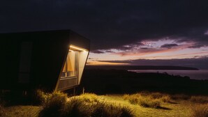 A modern, dark-toned building with a lit interior, situated in a grassy area with a view of a sunset over water. - CABN Kangaroo Island Ocean View Private Off Grid Luxury Accommodation (Willoughby)