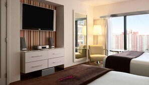 A hotel room with a TV, a desk with a computer, a chair, a bed, and a large window with a city view.
