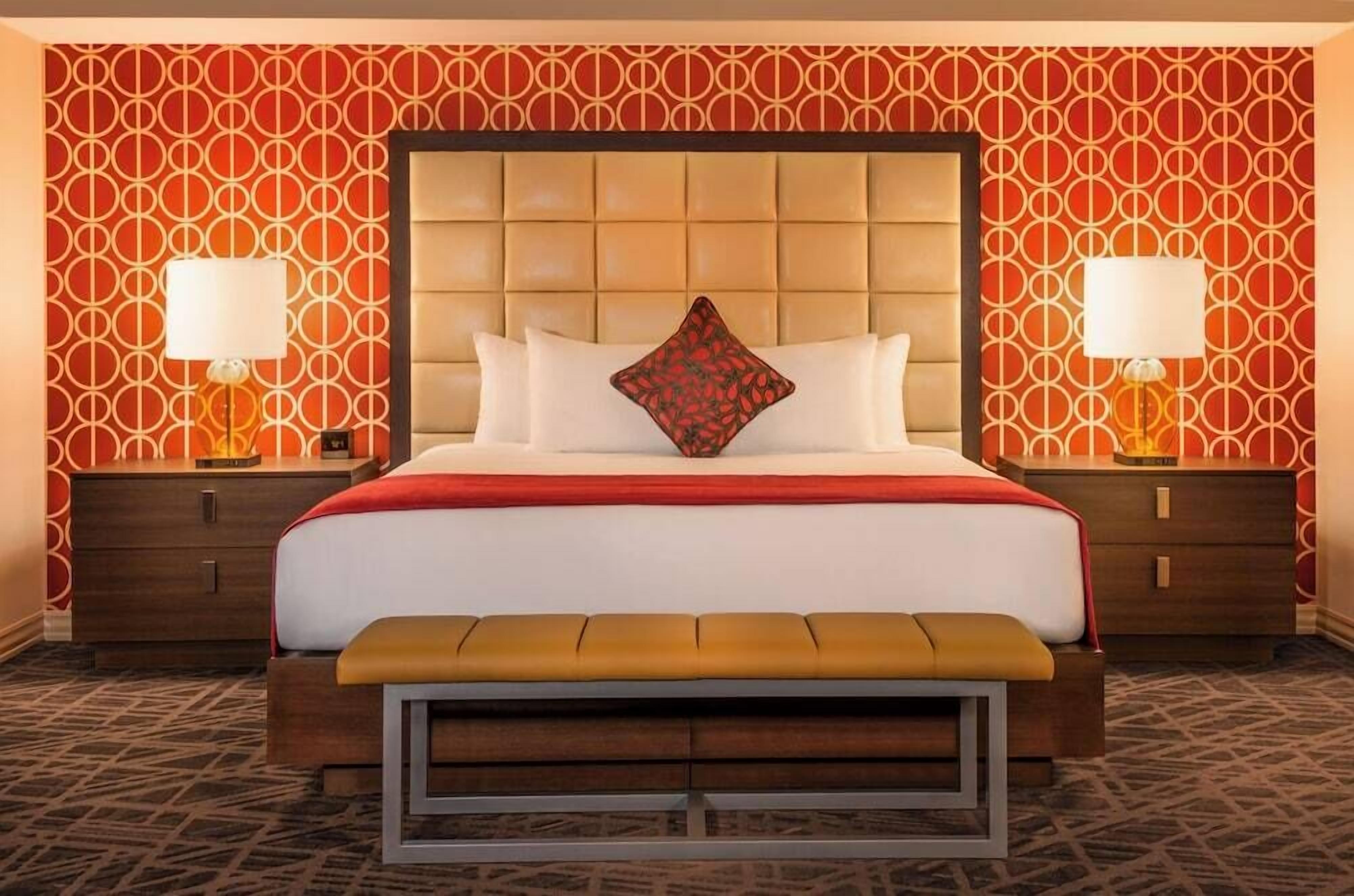 Une chambre d’hôtel avec un grand lit, deux tables de chevet avec des lampes, un banc au pied du lit et un papier peint à motifs.