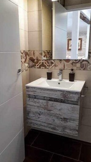 Een badkamer met een witte wasbak, twee donkere zeepdispensers en een spiegel boven de wasbak. De wanden zijn betegeld met een patroon.