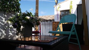 Eine Terrasse mit einem Korbstuhl-Tisch und -Stühlen, einem Blumentopf mit Blumen, einer Windmühle und einem Gebäude mit weißer Fassade und violetten Blumen.