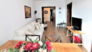 A living room with a white sofa, a dining table with a vase of flowers, a TV on a wooden stand, and a hallway leading to another room. - Casa Cervantes Toledo (Toledo)