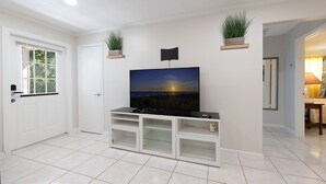 A living room with a flat-screen TV on a white entertainment unit, a door, and a window with blinds. - Let's Spend Some Time Together (Wellington)