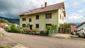 Uma casa de dois andares com telhado inclinado, várias janelas e um quintal cercado.
