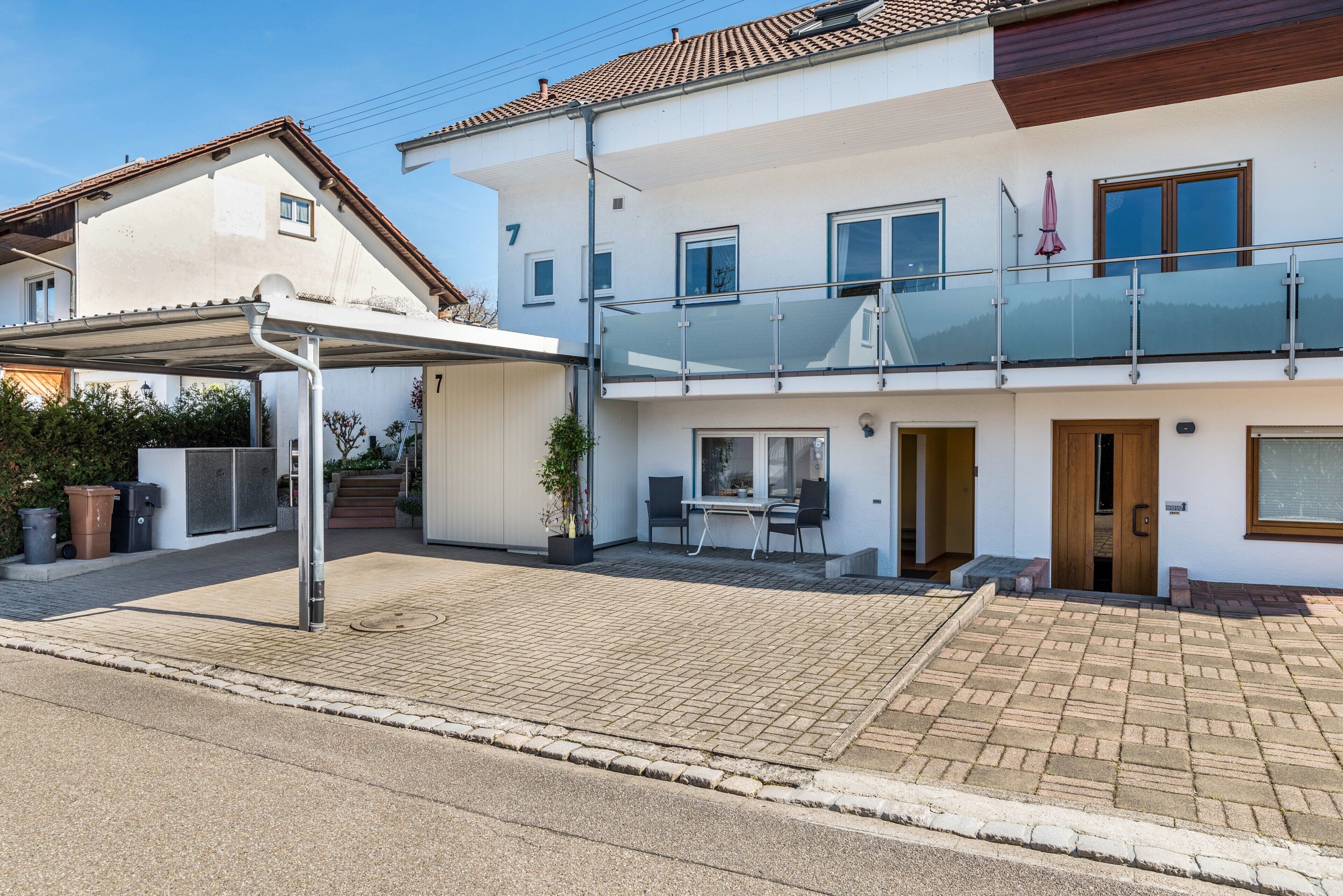Ein zweistöckiges Haus mit überdachter Auffahrt, Balkon und einer kleinen Außen-Sitzgelegenheit.