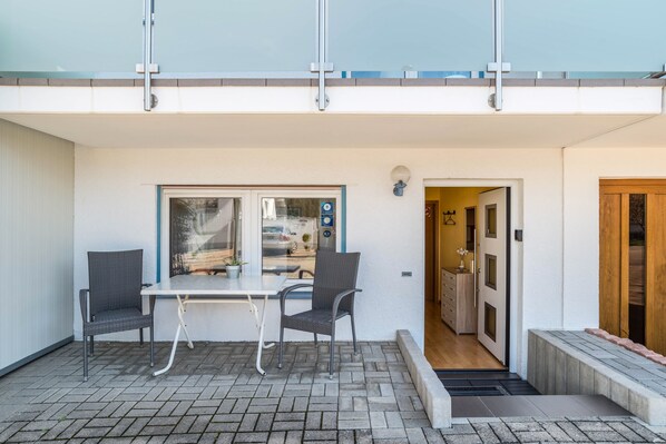A patio with a table and two chairs, a glass door leading to a hallway, and a wooden door on the right.