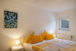 Une chambre à coucher comprenant un lit, des oreillers, une table de chevet, une lampe, une fenêtre et une œuvre d’art murale.