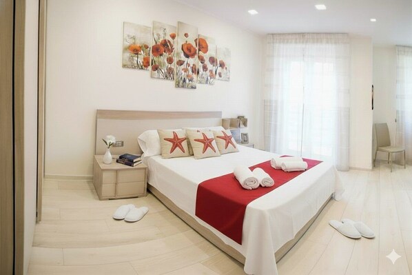 Habitación de hotel con una cama grande, una mesita de noche con lámpara, otra mesita y un ventanal con cortinas traslúcidas.