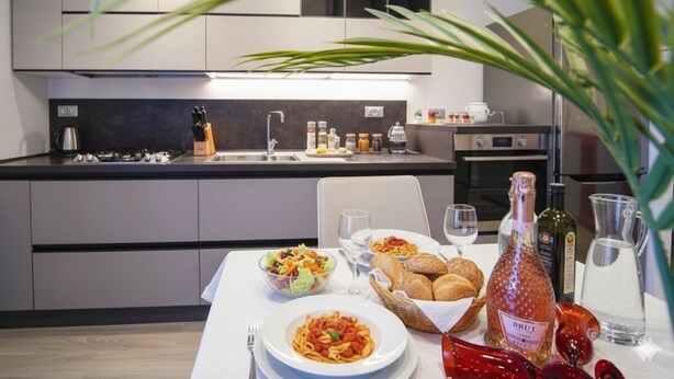 Una cocina moderna con una mesa de comedor preparada para una comida, incluyendo pasta, pan y una botella de vino Brut.