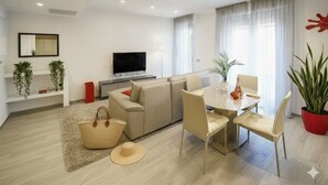 Una sala de estar moderna con un sofá, una mesa de comedor y sillas. Hay un televisor de pantalla plana en la pared, una lámpara y una planta en maceta.