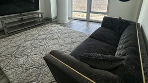 Una sala de estar con un televisor de pantalla plana grande, un sofá de color oscuro, un tapete estampado y un ventanal con vistas a un paisaje arbolado.