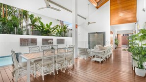 A spacious indoor area with a wooden floor, white wicker furniture, and a pool area visible through a glass wall. - 54 Ocean Drive Palm Cove (Palm Cove)