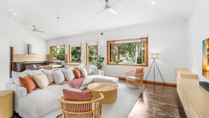 A spacious living room with a sofa, chairs, a round coffee table, and large windows offering a view of greenery. - 54 Ocean Drive Palm Cove (Palm Cove)